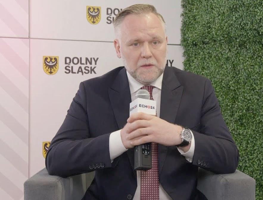 Dawid Jackiewicz podczas wystąpienia publicznego, z gestykulacją, na tle sali konferencyjnej.