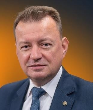 Mariusz Błaszczak na tle sprzętu wojskowego lub żołnierzy.