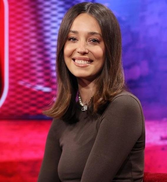 Izabella Krzan podczas prowadzenia programu telewizyjnego, dynamicznie gestykulująca, z mikrofonem w ręku, w tle elementy studia TV.